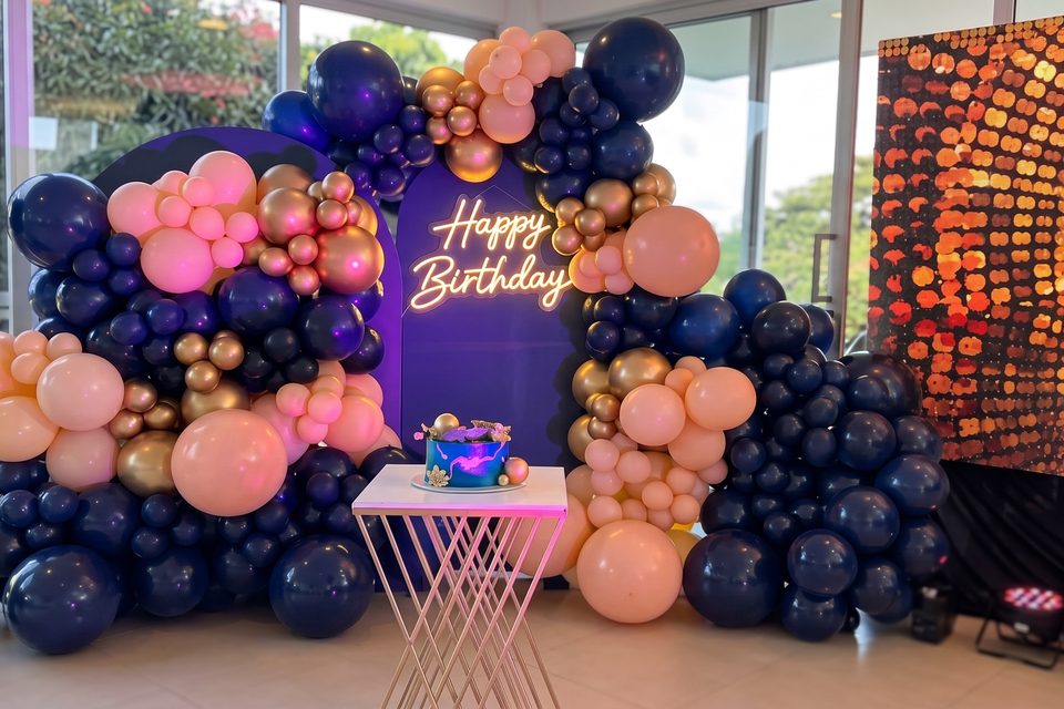 Arco de globos premium en azul, rosa y dorado alrededor de un letrero de neón Happy Birthday y la mesa de la torta